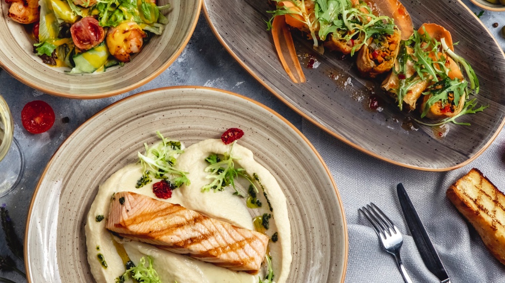 Three beautifully plated dishes – grilled salmon on a puree, shrimp salad, and vegetable rolls with arugula – showcasing how visually appealing digital menus lead to higher orders.